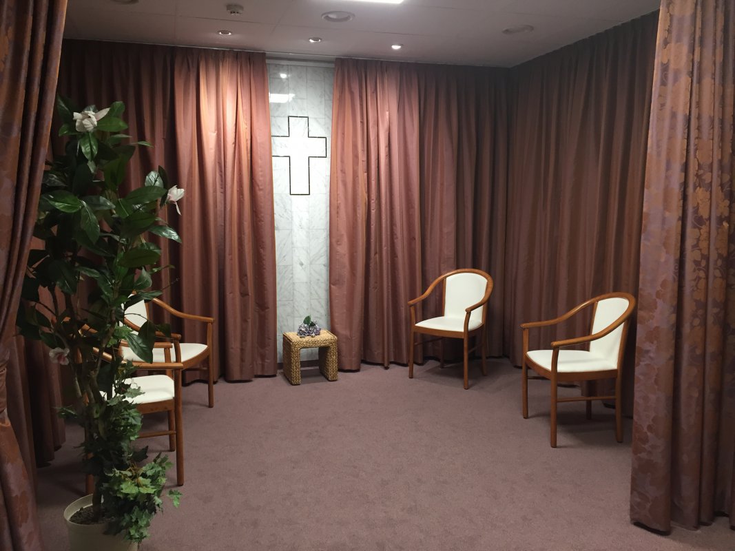 Blick in den "Raum der Stille". (c) Evangelisches Krankenhaus Ein Raum mit Teppich und warmen, gold-braunen bodenlangen Vorhängen ist zu sehen. Stühle, eine Pflanze und ein Kreuz an der Wand.